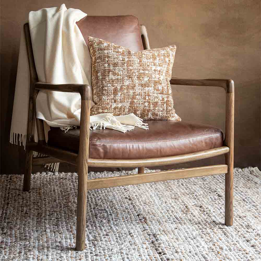 A brown leather armchair styled with a cream throw blanket draped over the side and a textured cream and brown textured pillow resting on the seat, set against a warm brown wall.