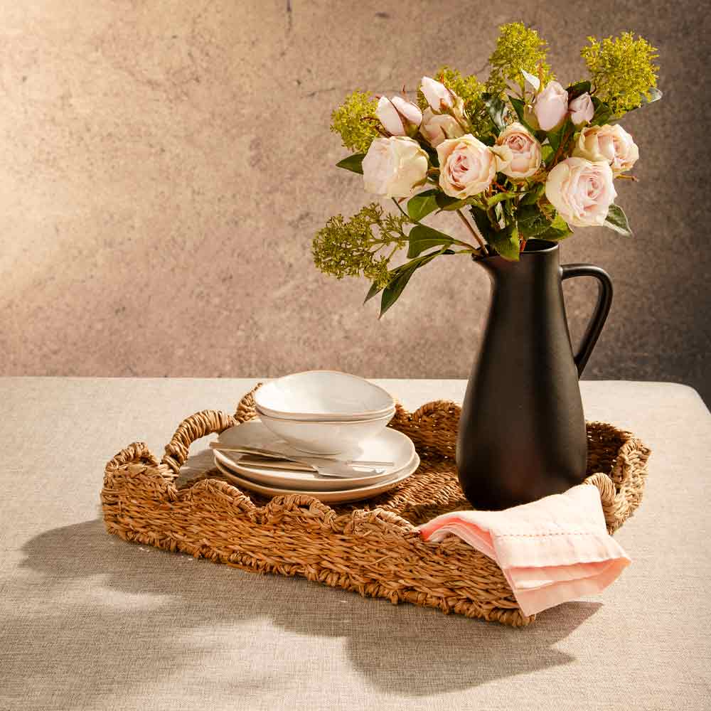 A styled tabletop scene with a scalloped woven rectangular tray holding a white dinnerware set and a folded pink napkin, next to a matte black pitcher filled with soft pink vintage rose spray and green skimmia foliage, against a textured beige wall.