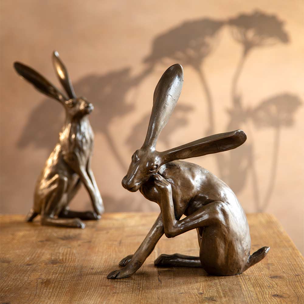 Two bronze-finished hare sculptures displayed on a wooden surface against a warm beige wall, with one in the foreground in a scratching pose and one in the background in a alert pose. Shadows of flower seed heads are cast on the wall.