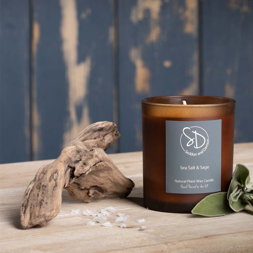 An amber glass jar candle labeled 'Sea Salt & Sage' by Seddon and Davison sits on a wooden surface beside driftwood, salt crystals, and green Sage leaves, with a distressed blue-grey wood plank background.