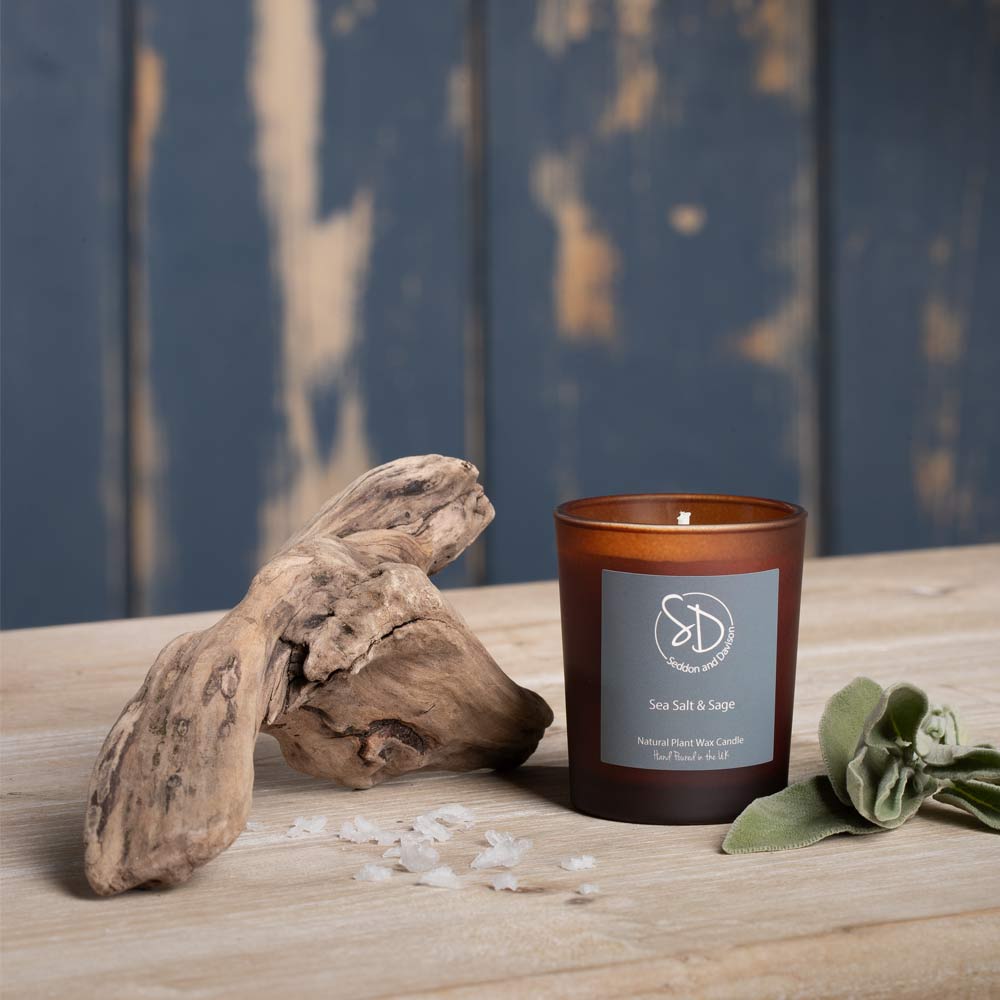 An amber glass Votive candle labeled 'Sea Salt & Sage' by Seddon and Davison sits on a light wood surface next to a piece of driftwood, scattered sea salt, and green Sage leaves, with a distressed blue wooden background.
