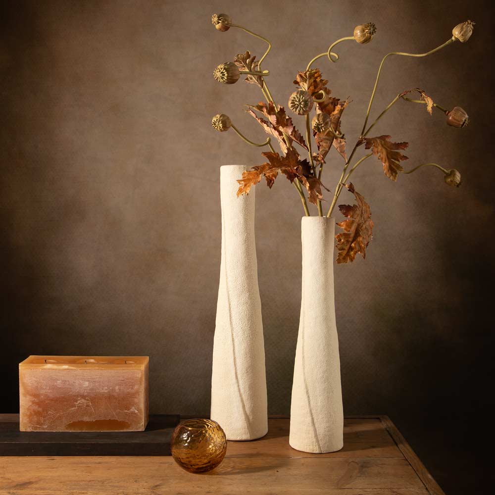 Two tall textured off-white ceramic vases one filled with faux stems brown leaves and seed pods, displayed on a wooden table against a dark brown background.