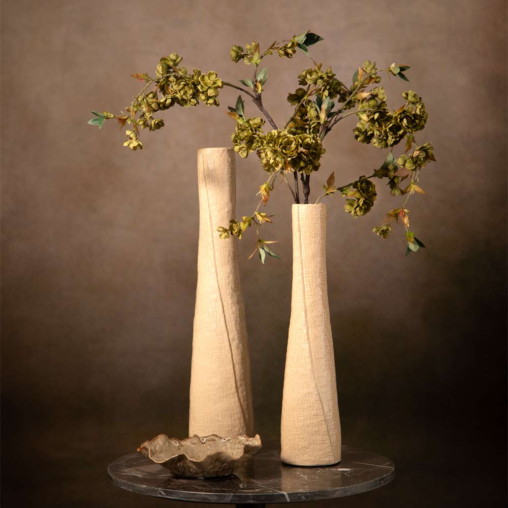 Two tall textured oatmeal vases on a round dark marble table, with faux green cherry blossom foliage arranged in the vase against a warm brown background.