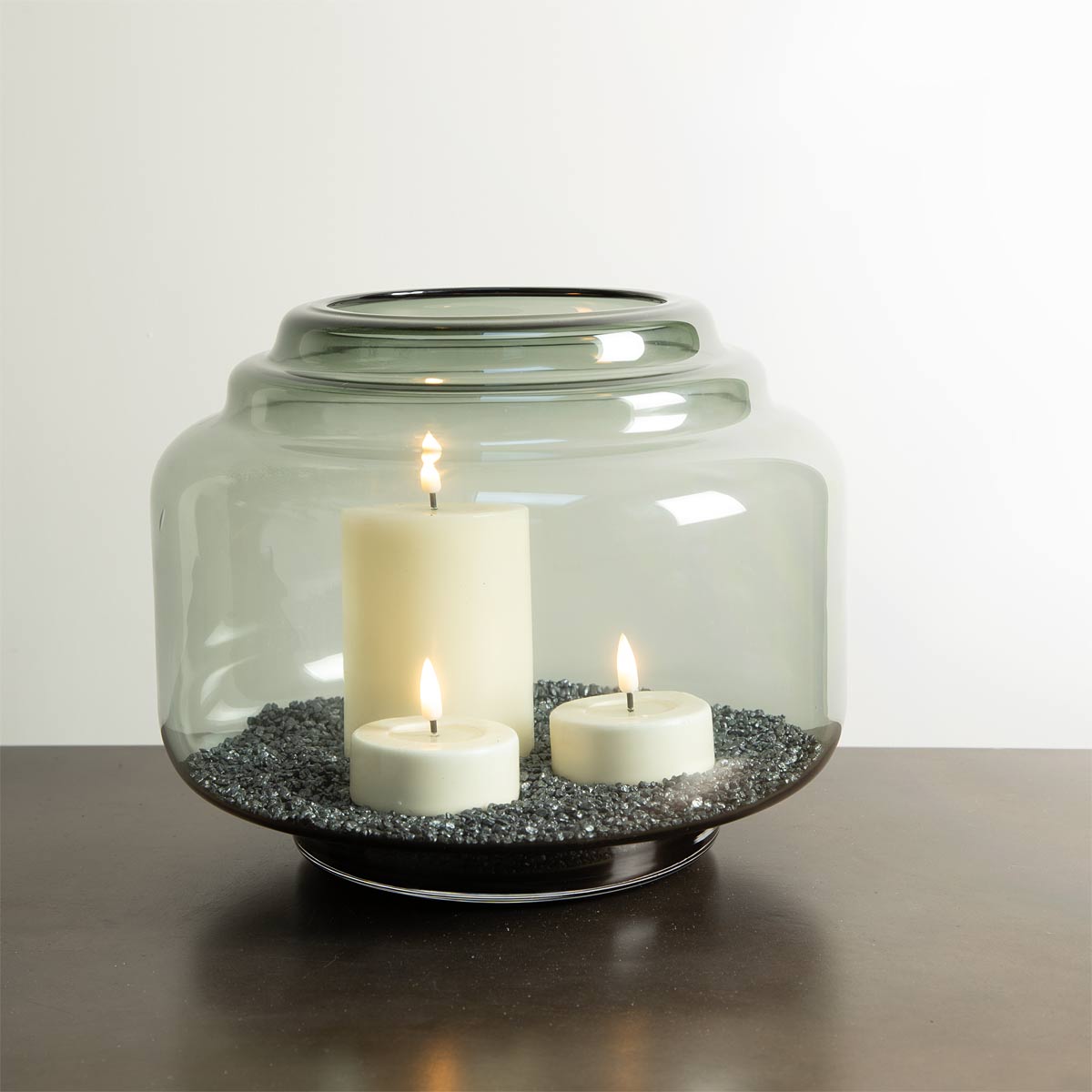 Wide smoked glass jar-style vase with black base, styled with pillar candles and decorative gravel inside.