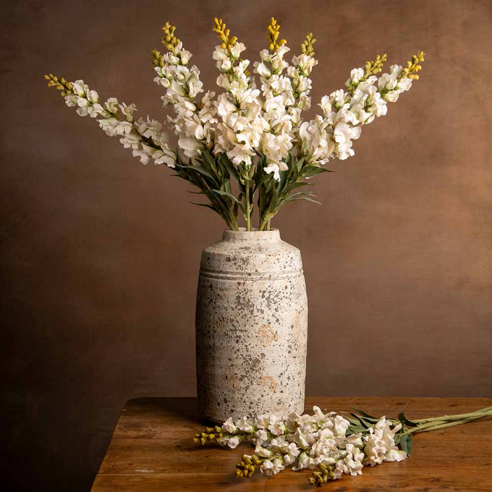 White artificial snapdragons with yellow accents arranged in a textured beige ceramic vase on a wooden table against a warm brown background.