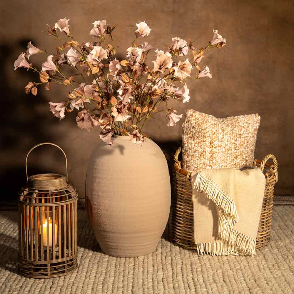 A styled interior scene with a large matte beige vase holding pale dusky pink flowers, a lit woven lantern on the left, and a wicker basket with a cream throw blanket and textured pillow on the right, set against a warm brown wall and woven rug.