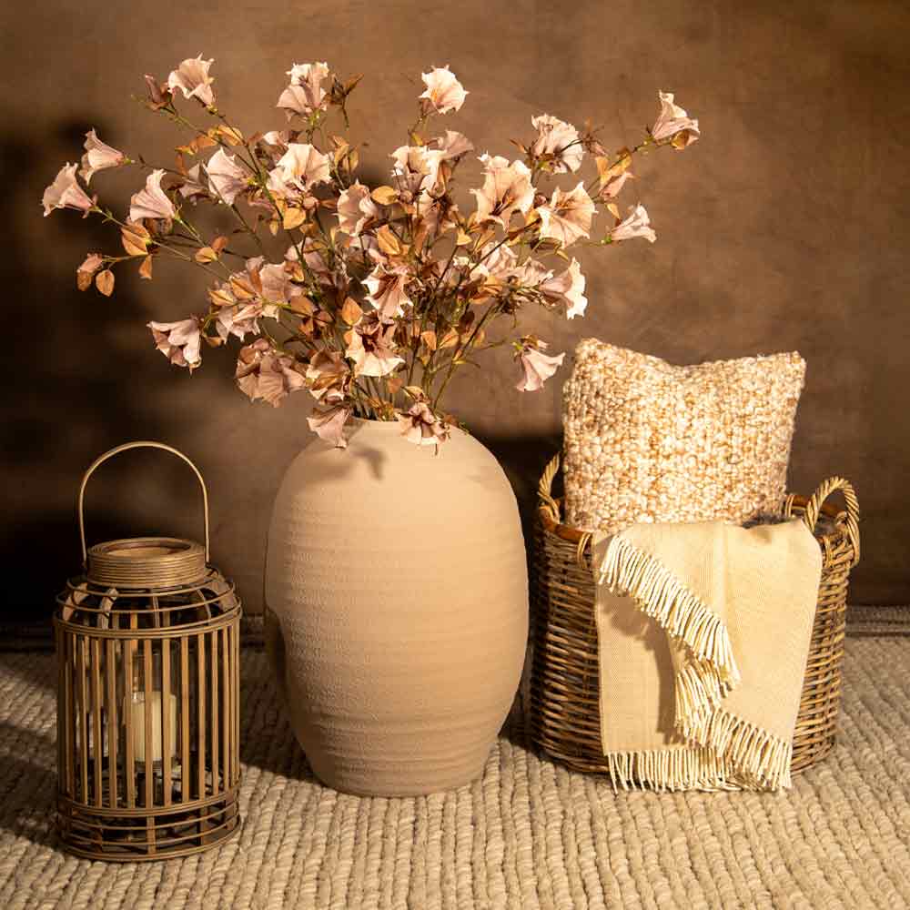 A styled home decor scene with a large matte beige ceramic vase holding pale blush pink flowers, a woven rattan basket with a cream throw blanket and textured pillow, and a lantern on a woven rug against a warm brown wall.