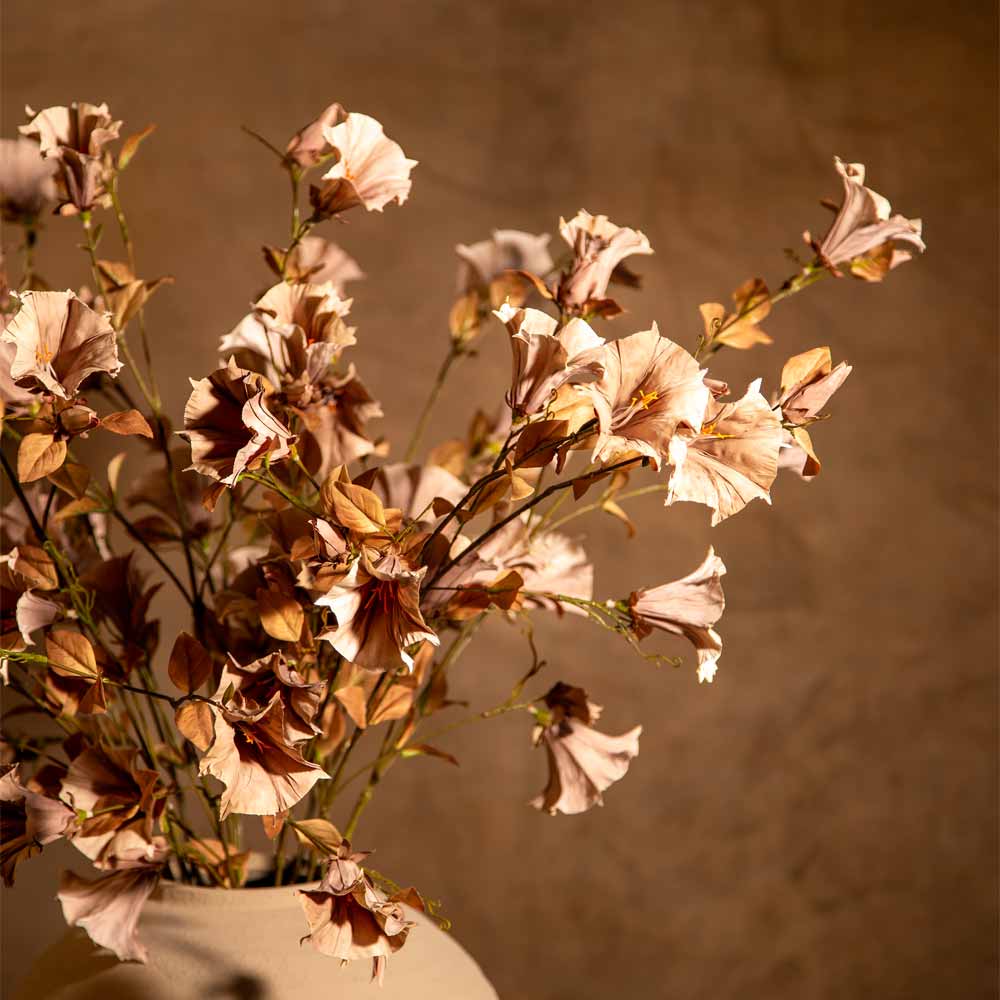 Artificial flowers in soft blush, pink , and brown tones arranged in a light beige vase against a warm brown textured background.