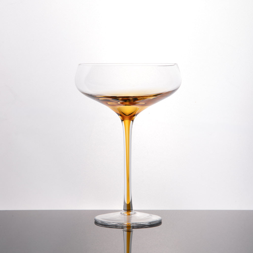 A clear coupe-style glass with a amber gradient at the base leading down the long stem and onto the round base, photographed on a dark reflective surface against a light gray studio background.