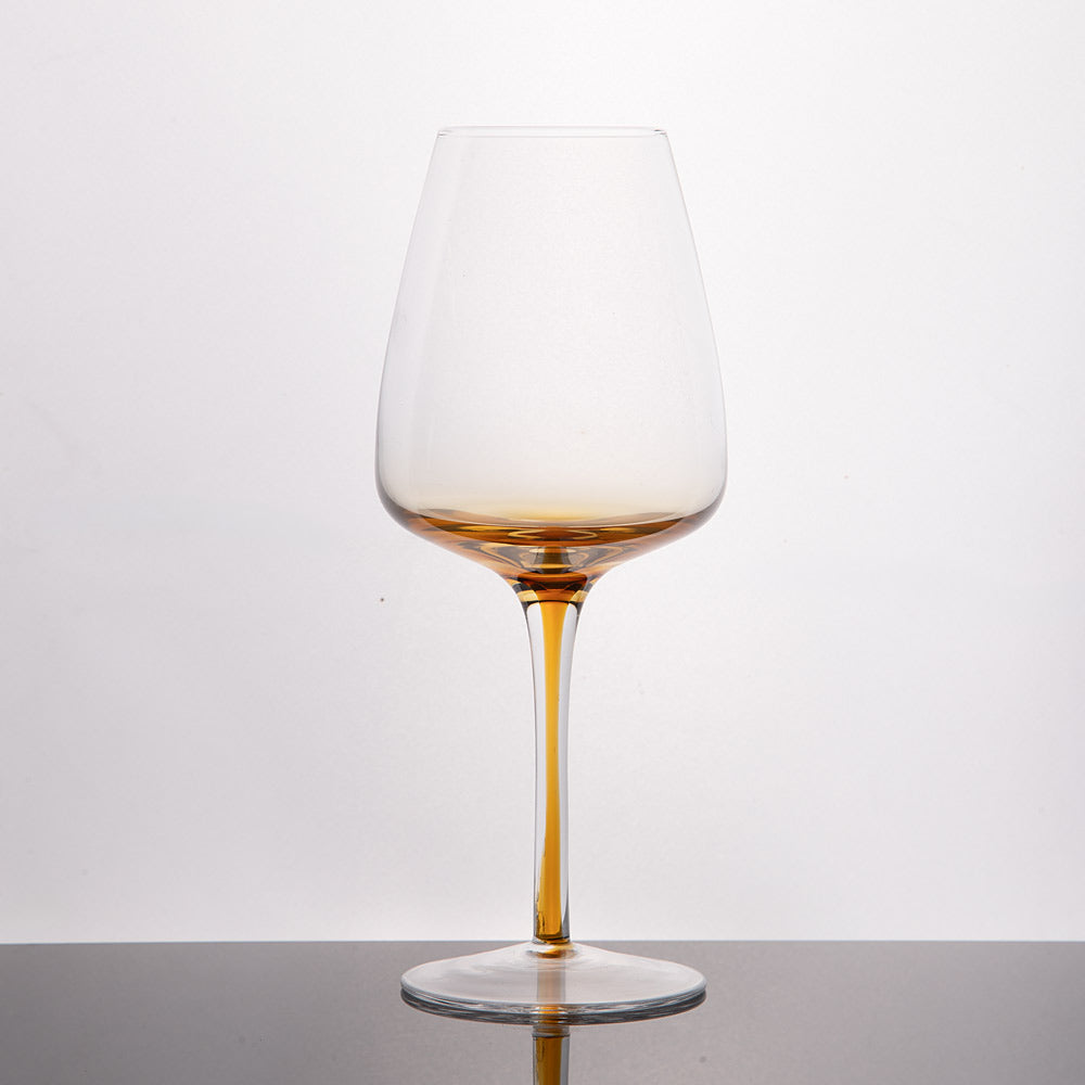 A clear wine glass with a distinctive amber-colored bottom and stem, standing on a dark reflective surface against a light gray background.