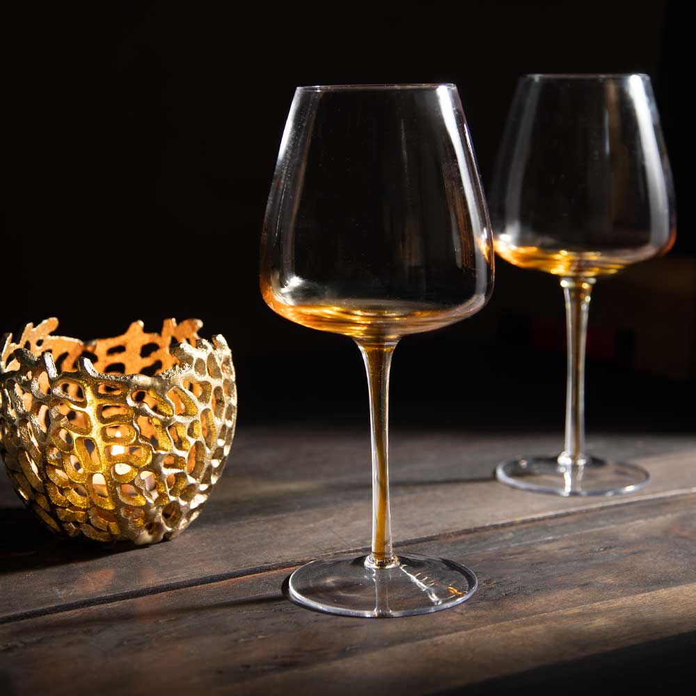 Two clear stemmed wine glasses with a clear wide bowl that grdients down to an amber stem, set on a dark wooden table next to a decorative gold candle holder against a black background.