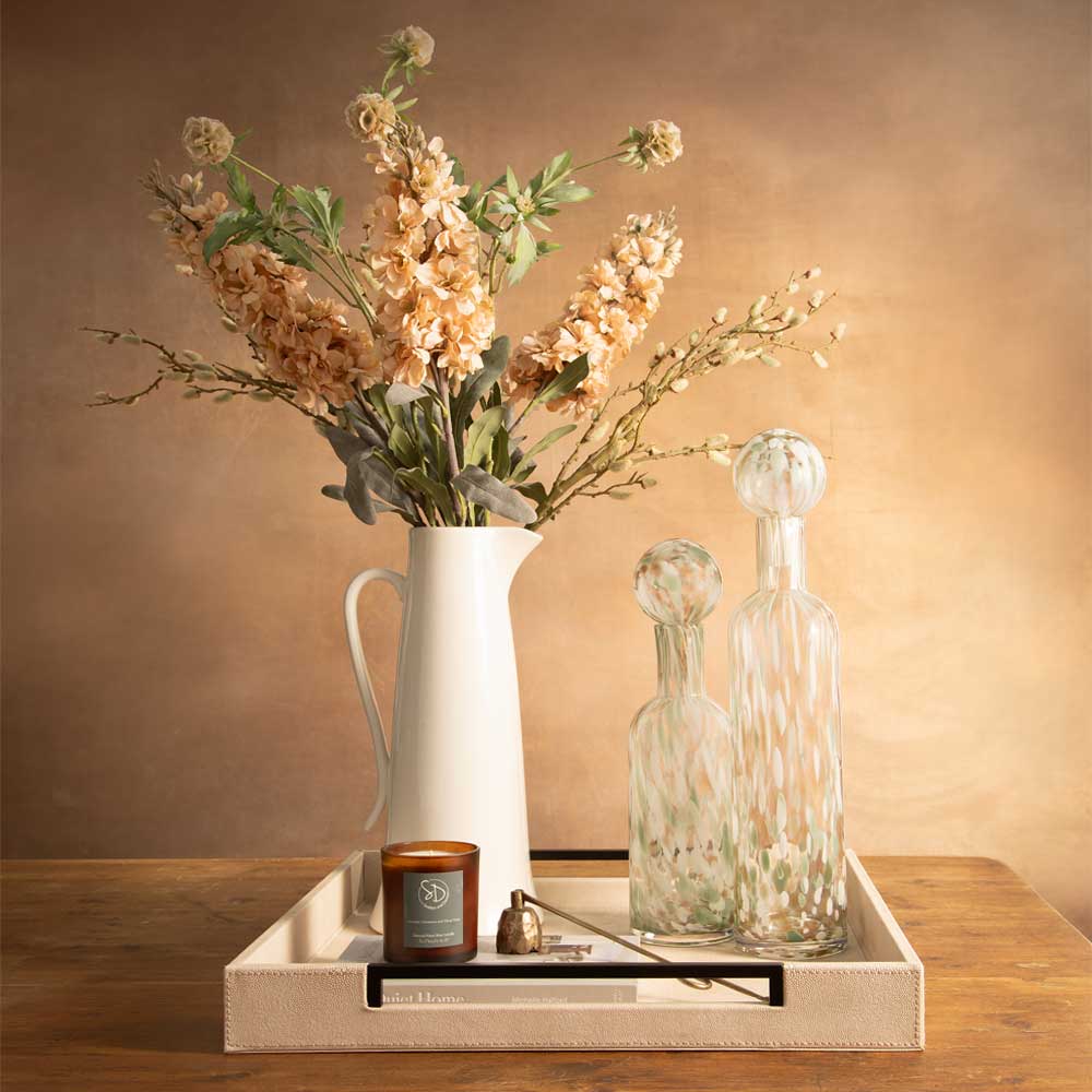 A styled tabletop arrangement featuring a tall ivory jug filled with dried cream and peach flowers, two clear textured glass decant bottles, a small amber candle, and a small brass bell resting on a rectangular tray against a warm brown backdrop.