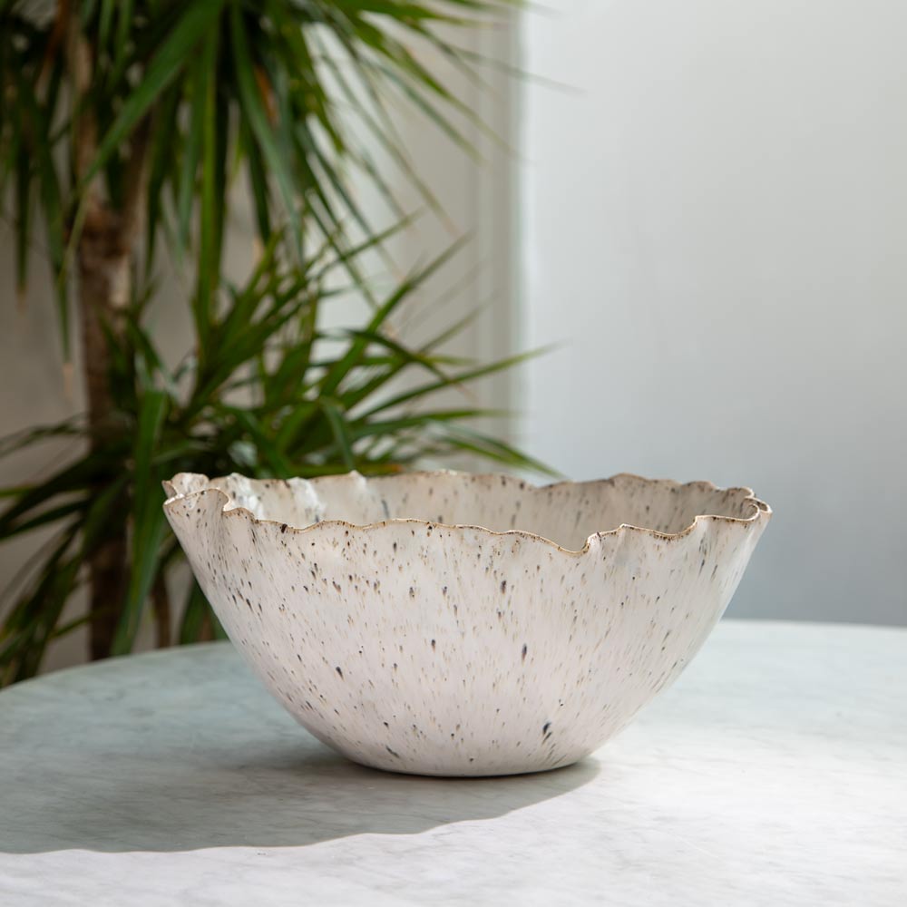 A ceramic bowl with a scalloped rim, speckled with dark brown, black and green flecks on a warm off-white glaze, set on a light grey marble surface with a blurred green plant in the background.