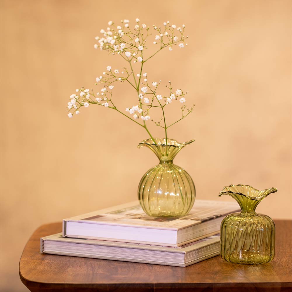 Two green glass vases with a single stem of flowers on top of books against a beige background