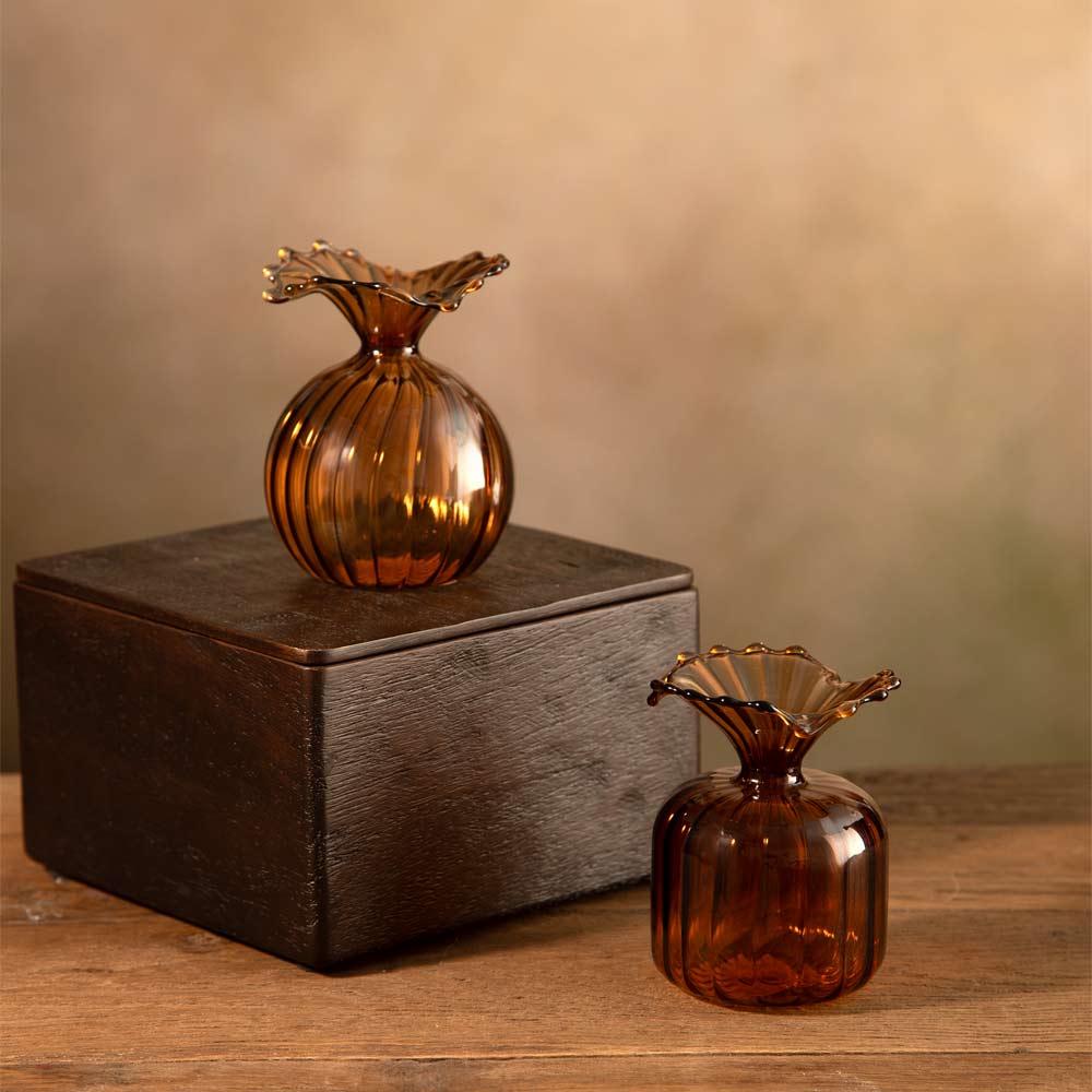 Two amber glass bud vase decor pieces with flared scalloped tops, one displayed on a dark wooden box and one standing beside it on a wooden surface against a warm beige background.