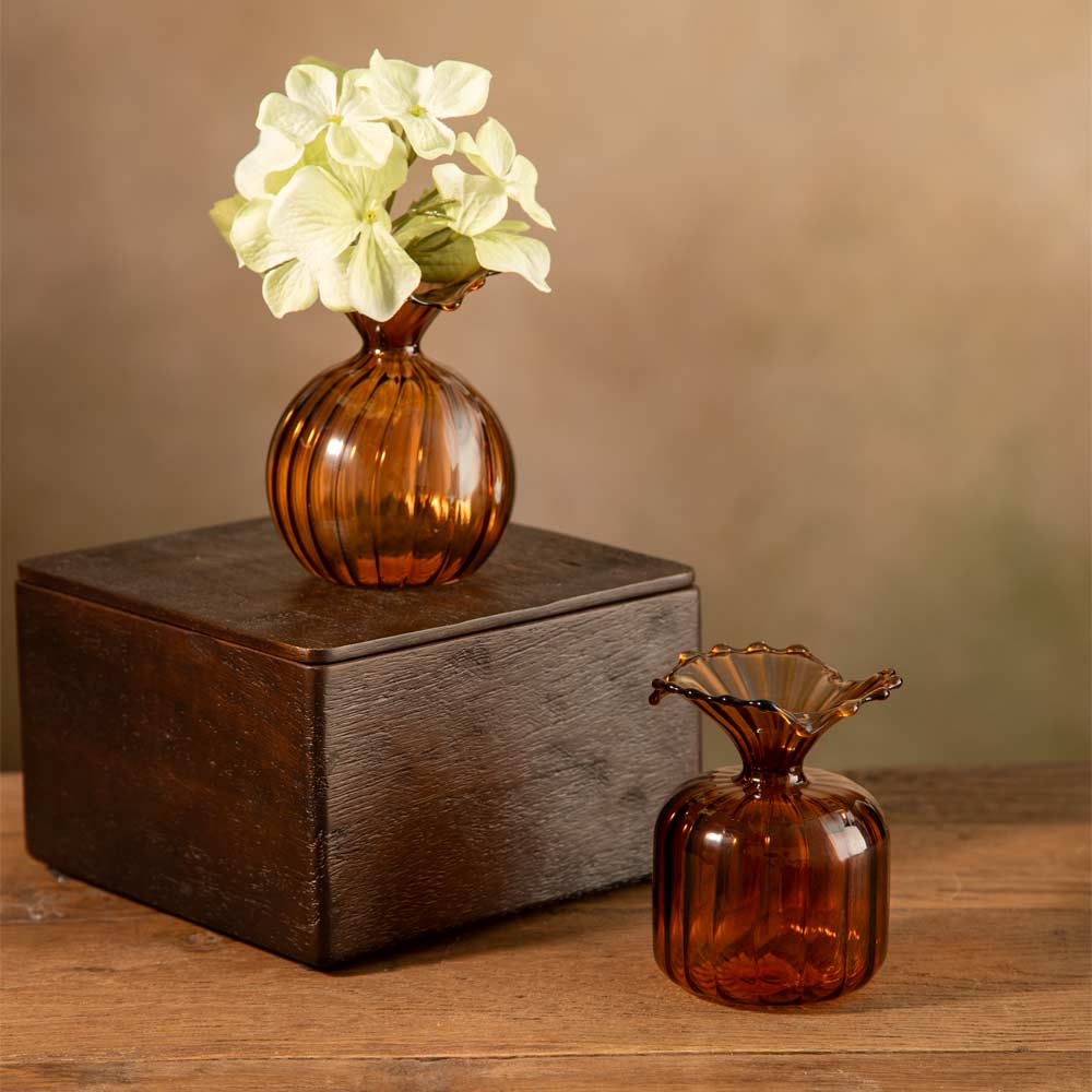 Two amber glass flower bud vases on a wooden table, with one vase filled with pale green faux hydrangea bloom and placed on top of a dark wooden box, and the other vase standing beside it.