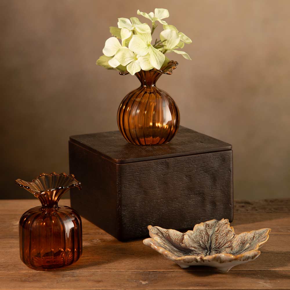 Two amber glass bud vase decor pieces on a wooden surface against a warm brown backdrop, with one holding pale green flowers and the other placed beside a dark rectangular box and a leaf-shaped dish.