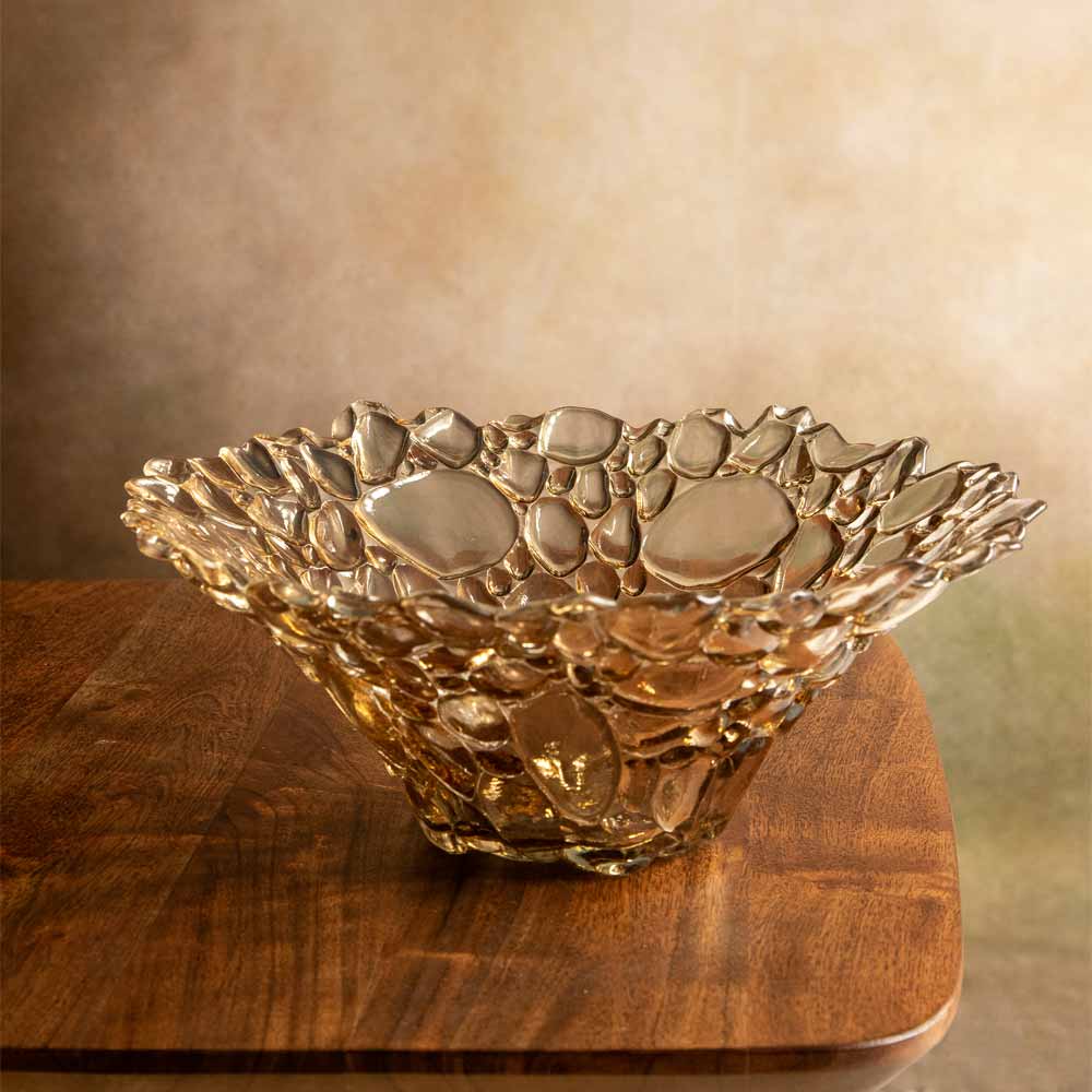 A decorative glass bowl with an organic, bubbly textured pattern, sitting on a wooden board against a warm, neutral background.