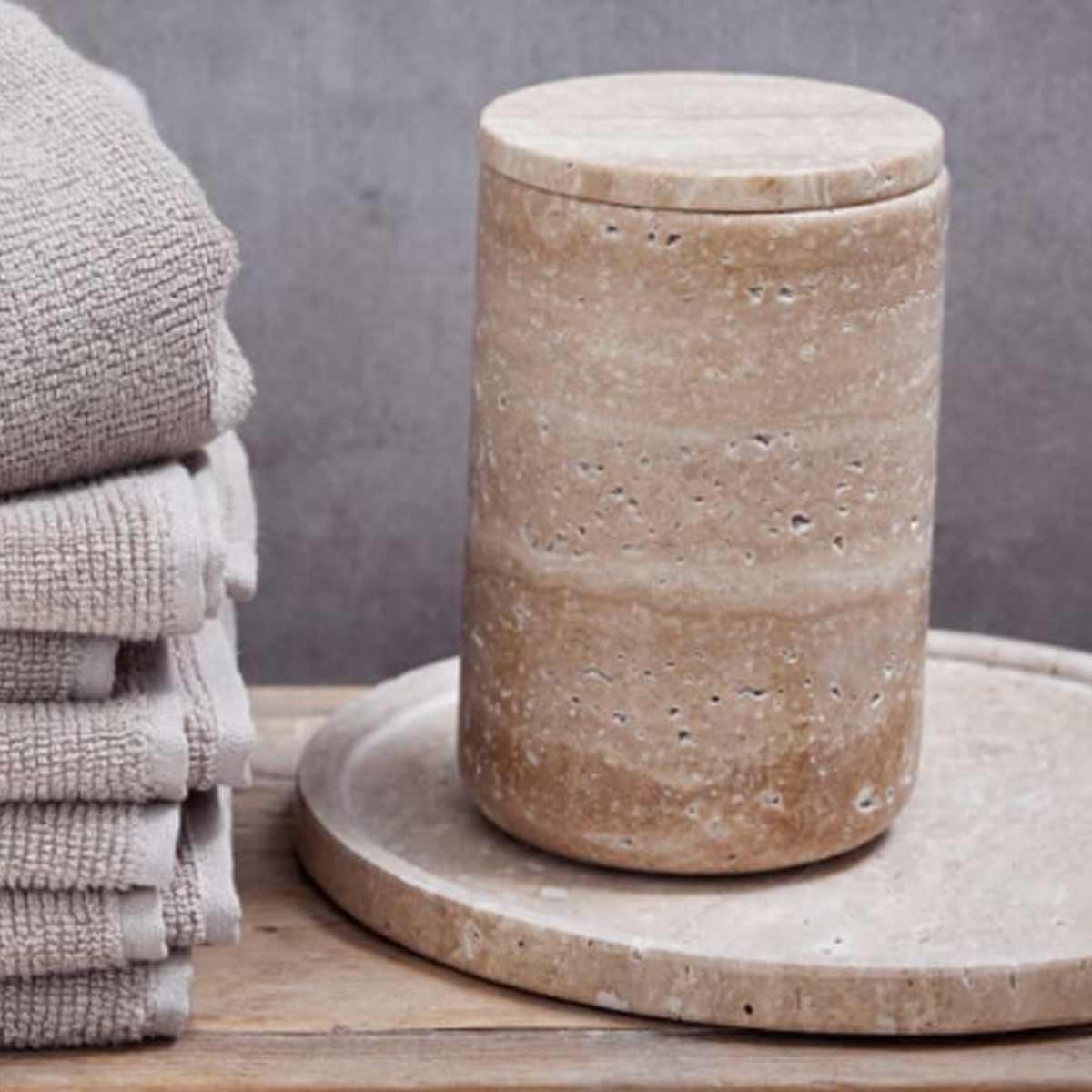 Travina Round Marble Tray in linen – a natural travertine tray with subtle stone variations, styled on a wooden surface with neatly folded towels and a matching travertine storage jar. A refined and practical piece for organising bathroom or home essentials.