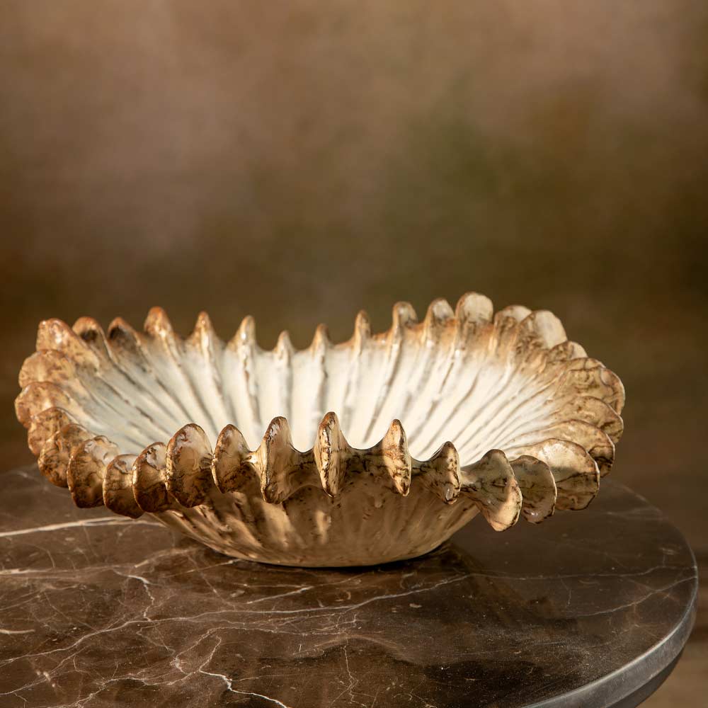 A decorative ceramic shell like bowl with a wide, flared scalloped rim, shown on a dark marble surface against a warm brown studio background.