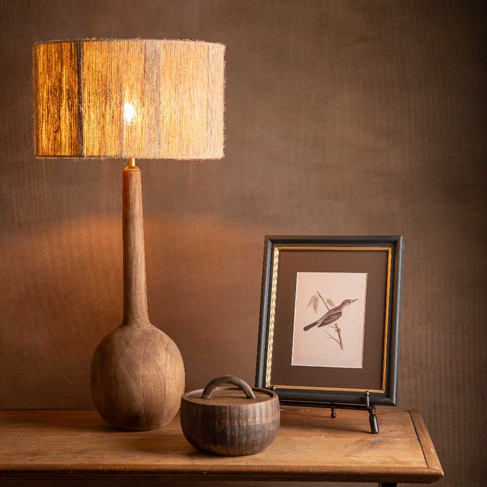 A rustic wooden table lamp with a textured beige and brown fringed drum shade and a rounded wooden base, shown lit on a wooden table next to a framed bird print and a small wooden bowl.