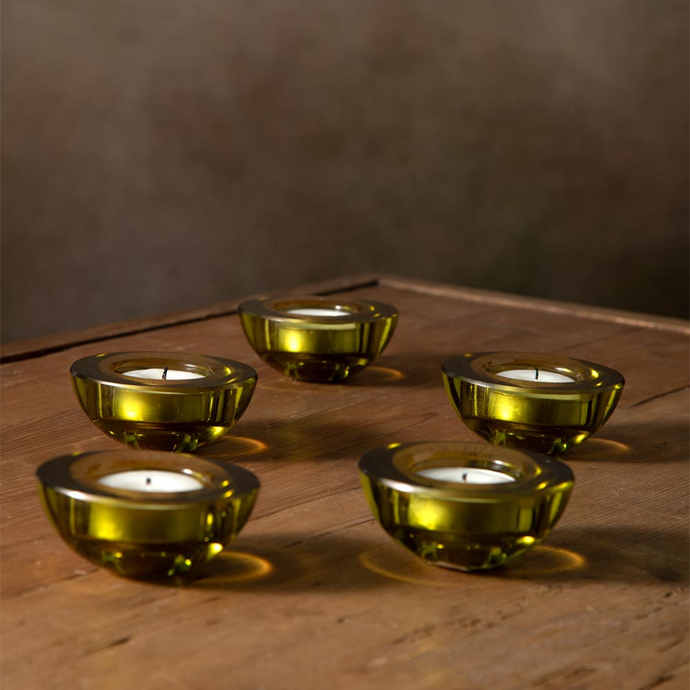 Five small green glass tealight candle holders arranged on a rustic wooden surface against a dark brown wall background.