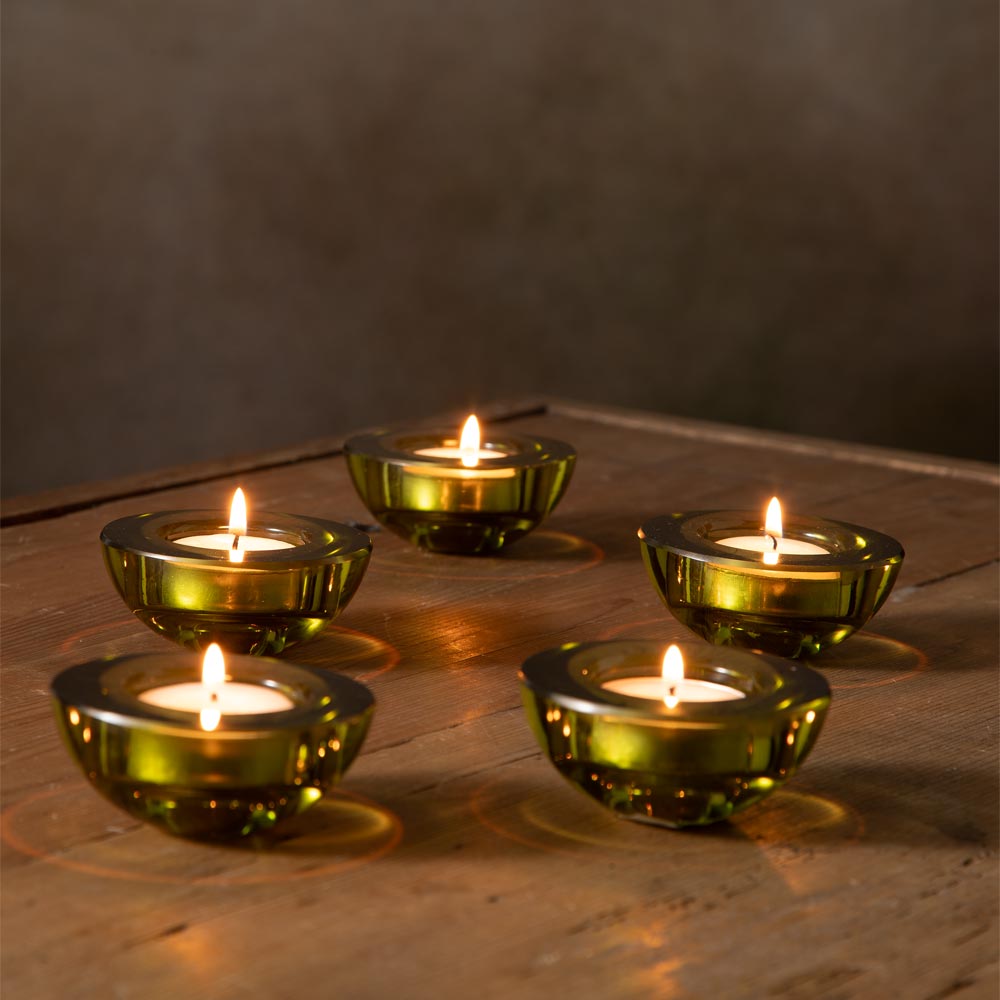Five small green glass tealight candle holders with lit candles, arranged on a rustic wooden surface against a dark brown wall background.
