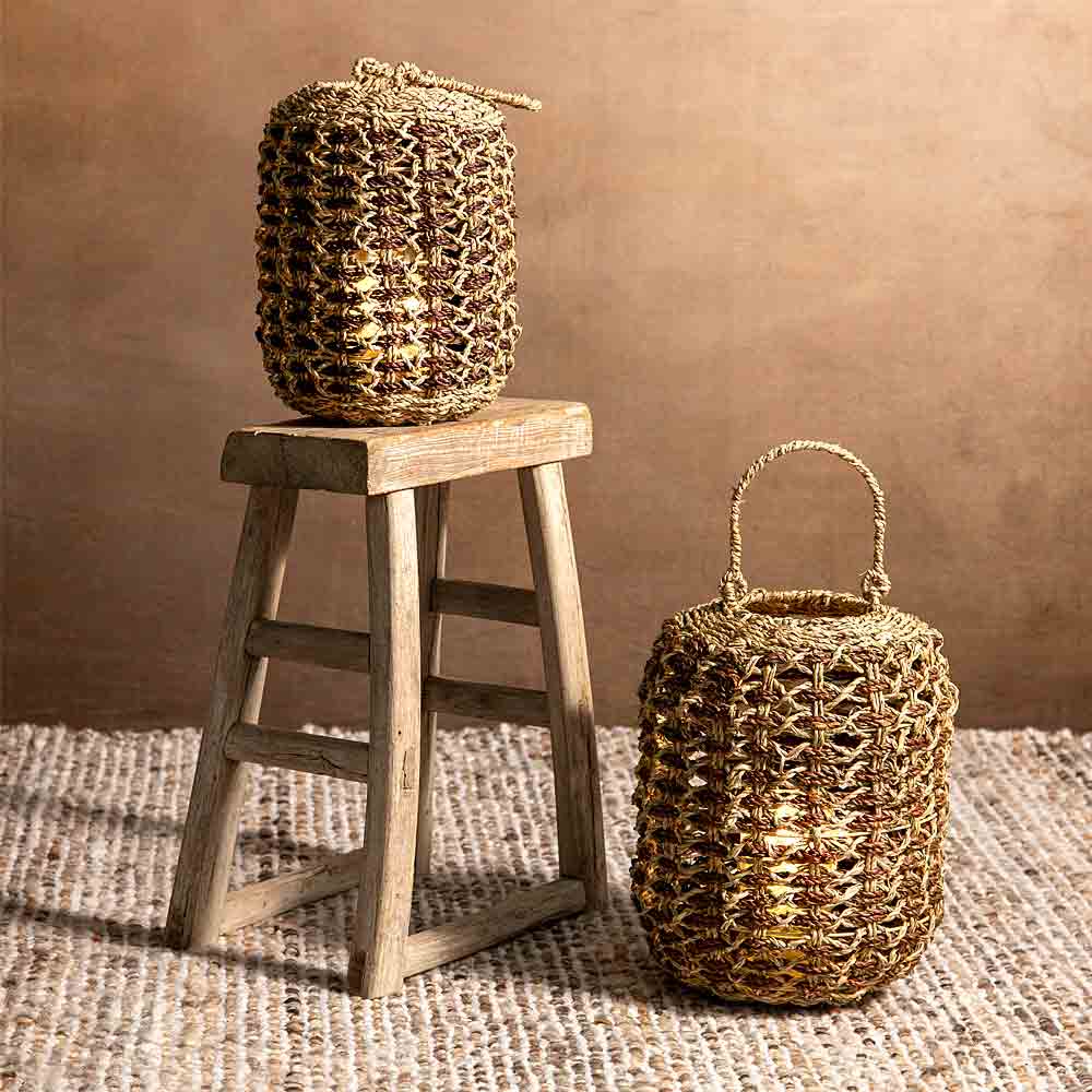 Two woven natural-fiber baskets with handles, one displayed on a rustic wooden stool and the other on a woven rug against a warm brown backdrop.