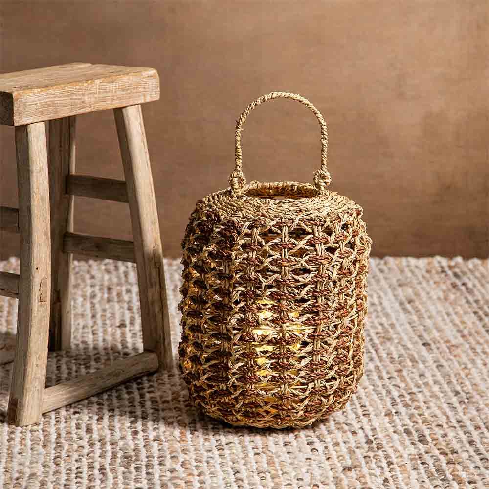 A woven natural-fiber basket bag with a top handle, styled next to a rustic wooden stool on a textured woven rug against a warm brown background.