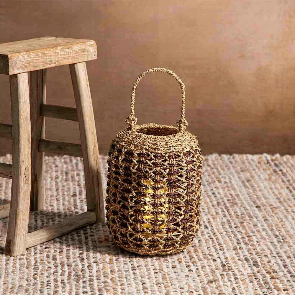 A small woven basket bag with a top handle, made from natural tan and dark brown raffia, styled next to a rustic wooden stool on a textured woven rug against a warm brown wall.