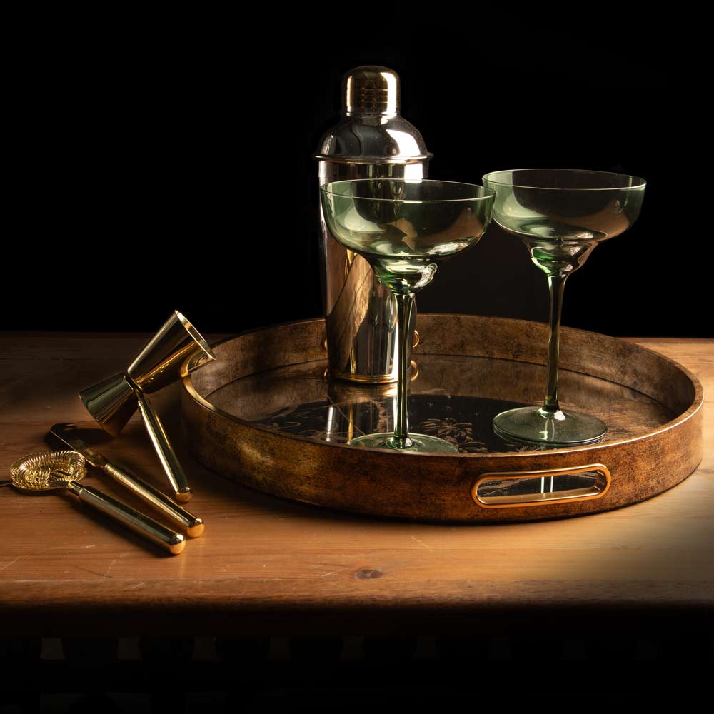 A bar-themed still life featuring a round gold - toned tray holding a two tone metal shaker and two green coupe glasses, with brass-toned cocktail tools resting on a wooden surface against a black background.