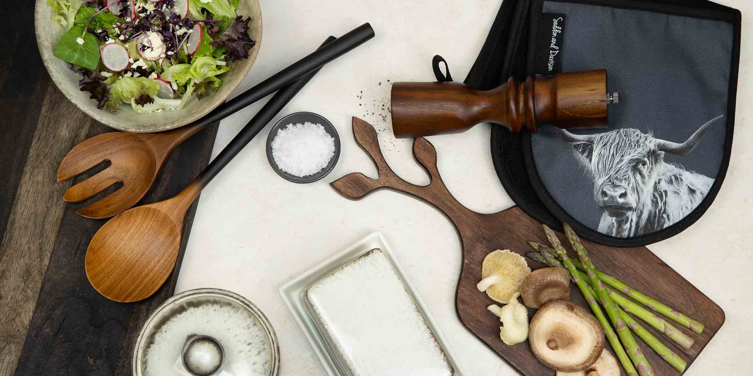 Kitchen serveware with wooden salad spoons, fresh salad, salt, vegetables, and Highland cow oven gloves.