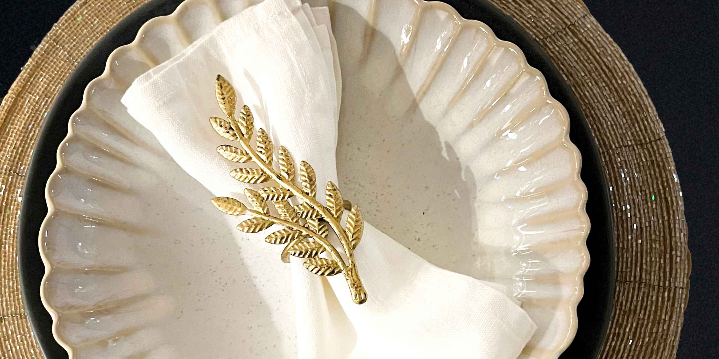 Elegant table setting featuring a white scalloped ceramic plate, gold beaded placemat, and a gold olive branch napkin ring on a folded white linen napkin.