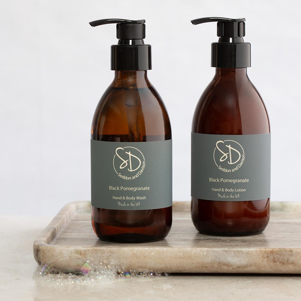 Two amber glass pump bottles with black dispensers and grey labels, shown side by side on a light stone tray. The labels read Seddon and Davison  Black Pomegranate Hand & Body Wash and Hand & Body Lotion.