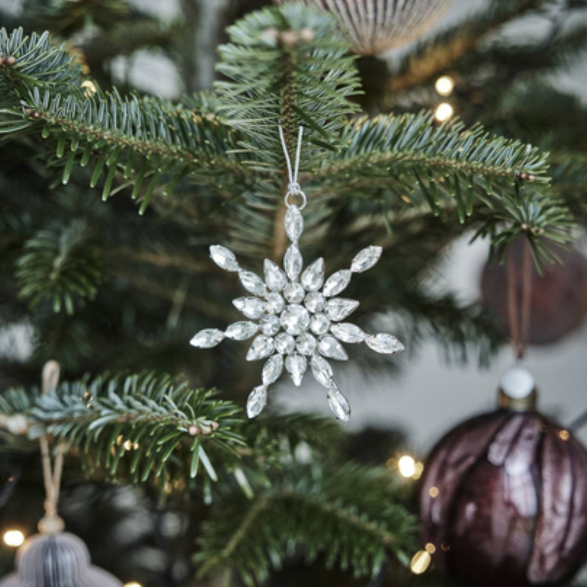 Decorative Ada Festive Silver Glass Tree Snowflake Tree Ornament with clear gemstones on a Christmas tree with blurred lights in the background