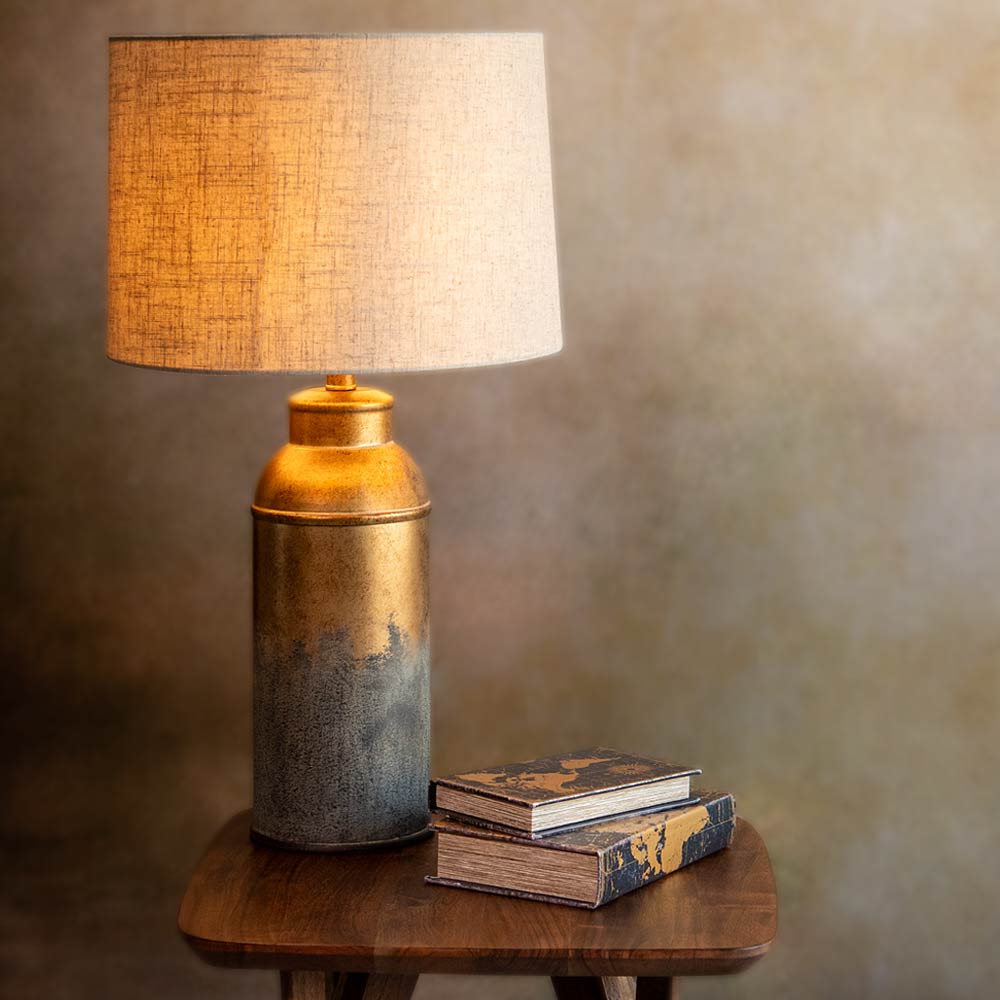 A table lamp with a textured beige drum shade and a metallic cylindrical base that transitions from gold at the top to dark Brown - grey petina at the bottom, shown lit on a wooden table next to two stacked books against a warm, textured grey -brown background.
