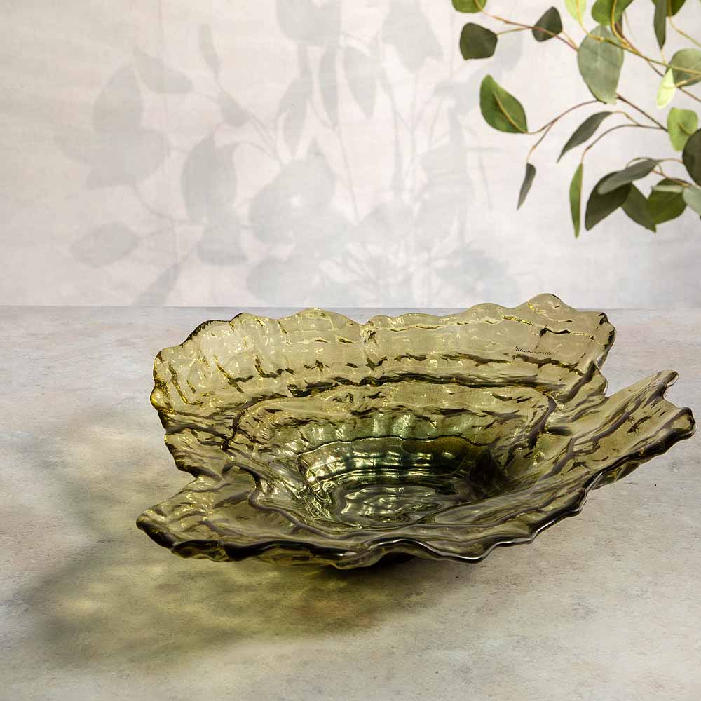 A textured, organic-shaped glass bowl in a muted olive green color, styled on a light gray surface with soft shadows and eucalyptus leaves in the background.
