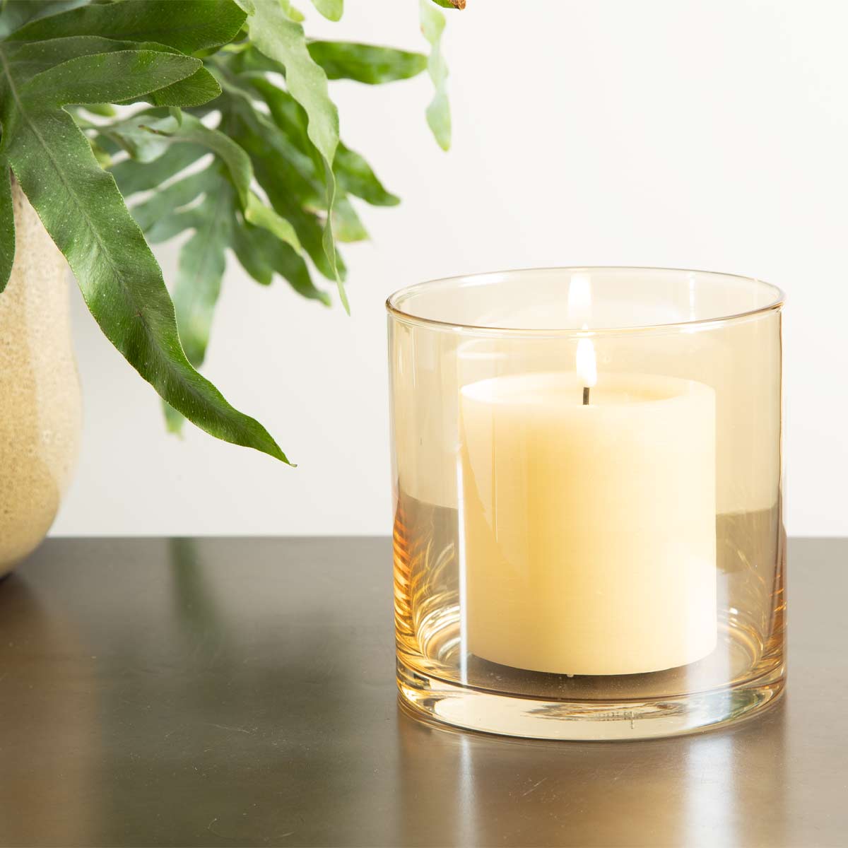 Amber glass hurricane candle holder with pillar candle on dark tabletop, styled beside green foliage

