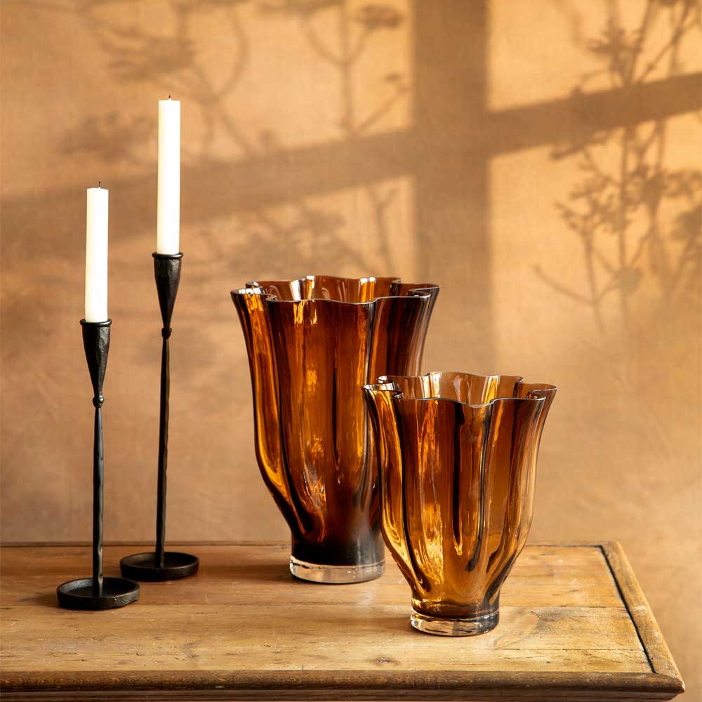 Two amber-brown translucent glass vases with flared wave scalloped rims sit on a wooden table, alongside two black candlesticks with white candles, against a warm beige wall with soft shadow patterns.