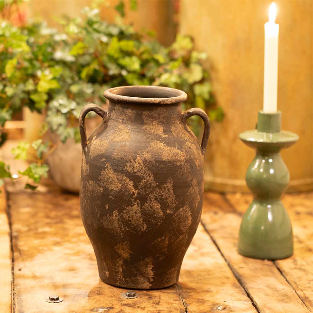 Vintage-style Annchi Brown Terracotta Vase on a wooden surface with a green candlestick and plants in the background.