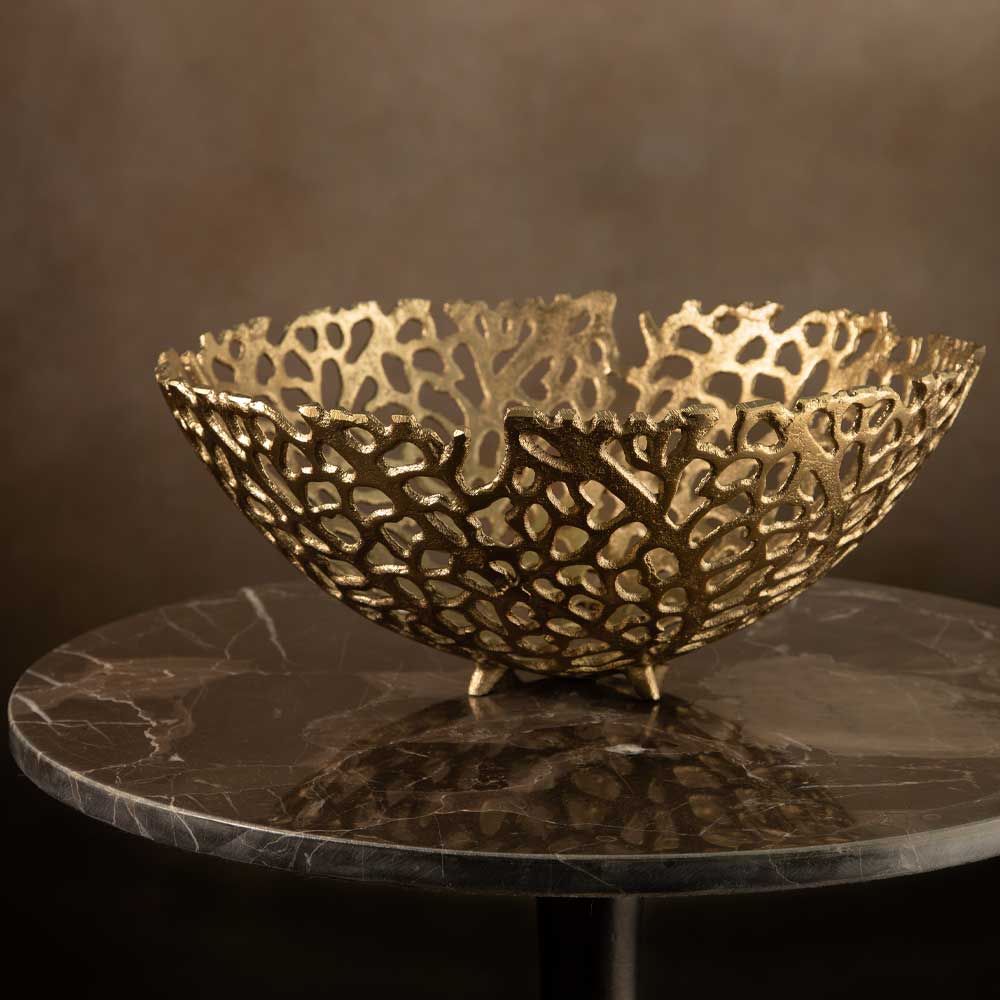 A decorative gold-toned metal bowl with an intricate openwork leaf or coral-like pattern, displayed on a round dark marble table against a warm brown background.