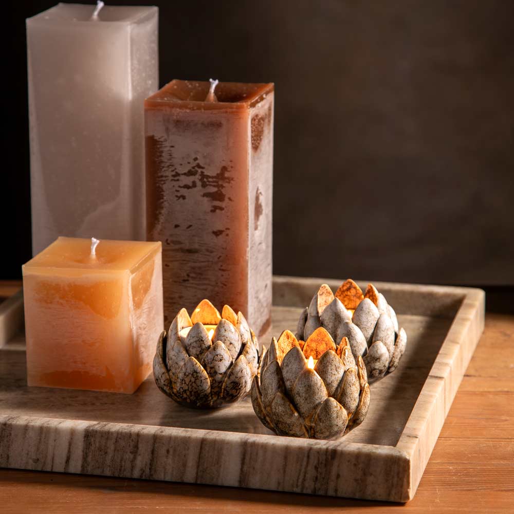 Three artichoke shaped decorative tealight holders  with lit wicks glowing orange arranged on a square  marble tray, including within the scene there are tall rectangular pillar candles in frosted off-white,  mottled brown and the shorter one in a  square caramel colour.