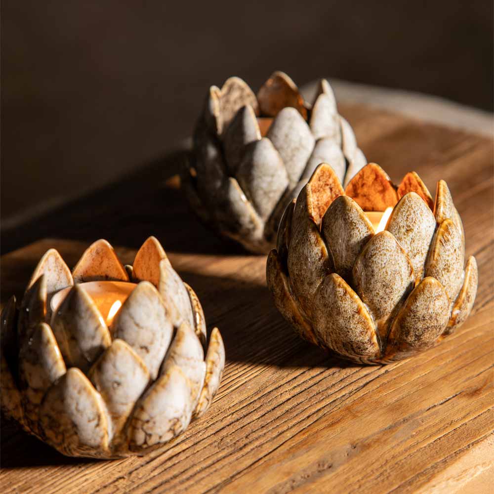 Three artichoke-shaped ceramic candle holders with a rustic, weathered finish, each containing a glowing tea light, arranged on a wooden surface.