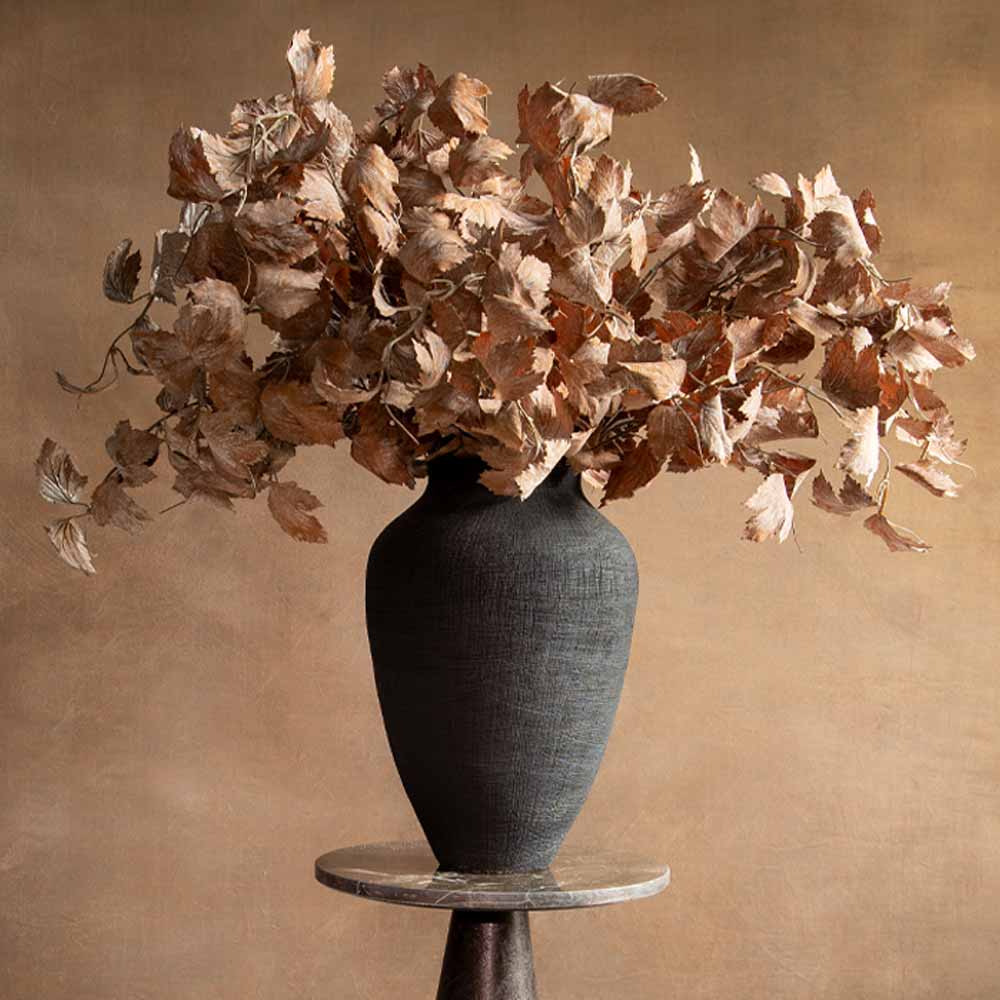 A Faux dried vine leaves arrangement in a black textured ceramic vase, displayed on a round marble topped pedestal against a warm brown wall.