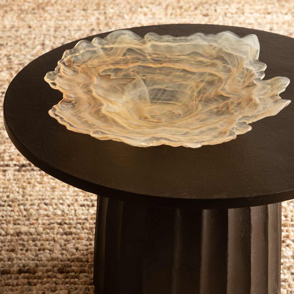 A translucent amber and cream Glass bowl with organic wavy edges, displayed on a round black side table over a beige and brown textured rug.