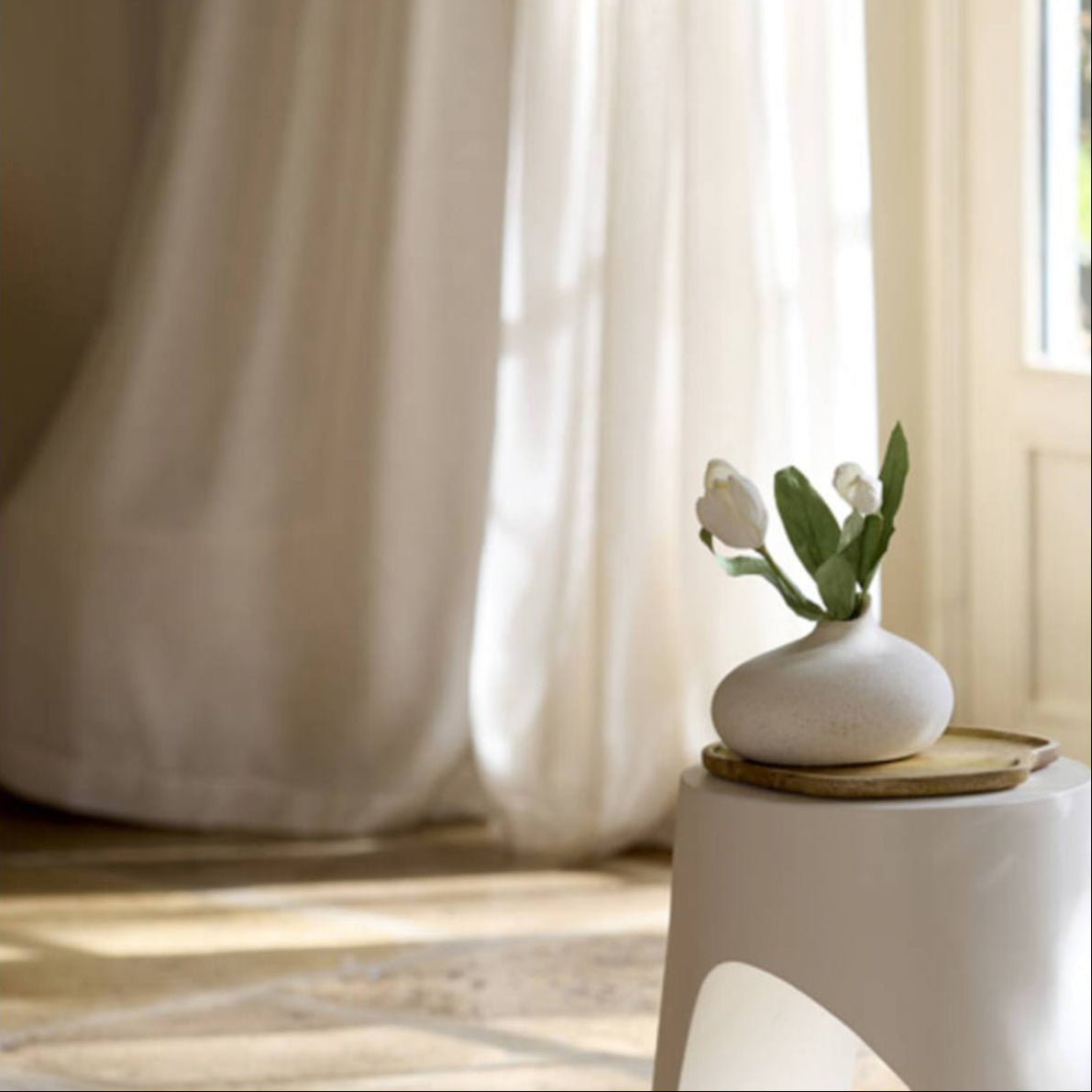White Assaga Cream Ceramic Short Vase with flowers on a white stool in front of sheer curtains