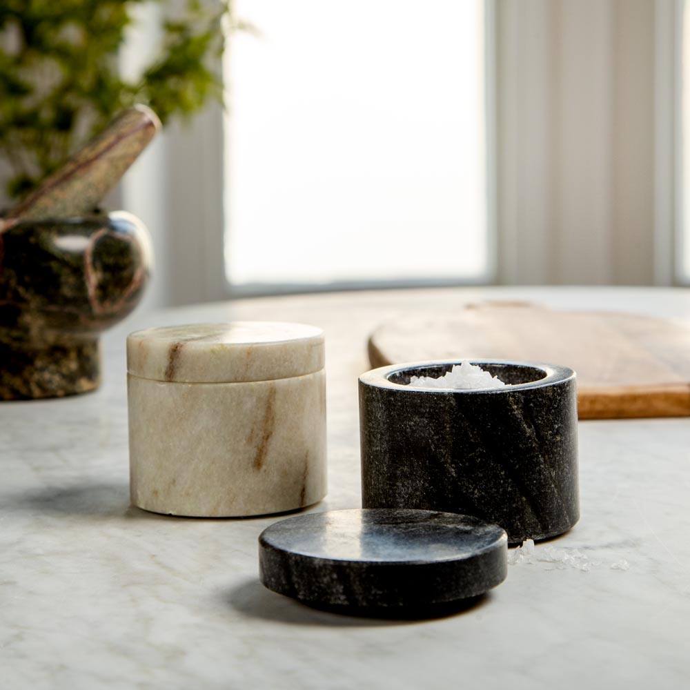 Two round marble salt and pepper containers on a light marble countertop, with one container open showing coarse salt and its lid placed in front. A blurred kitchen background with a plant and wooden board is visible.