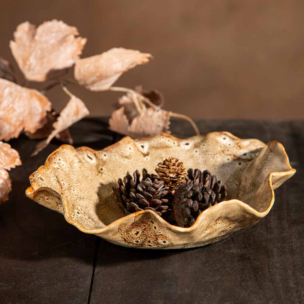A rustic, organic - shaped ceramic bowl with a textured, speckled beige and brown glaze, holding pinecones on a dark wooden surface with faux leaves in the background.