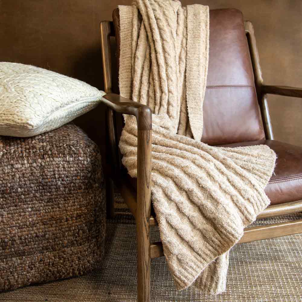 A chunky knit throw blanket in a warm beige color is draped over a mid-century modern wooden armchair with brown leather upholstery, next to a woven pouf and a textured pillow.