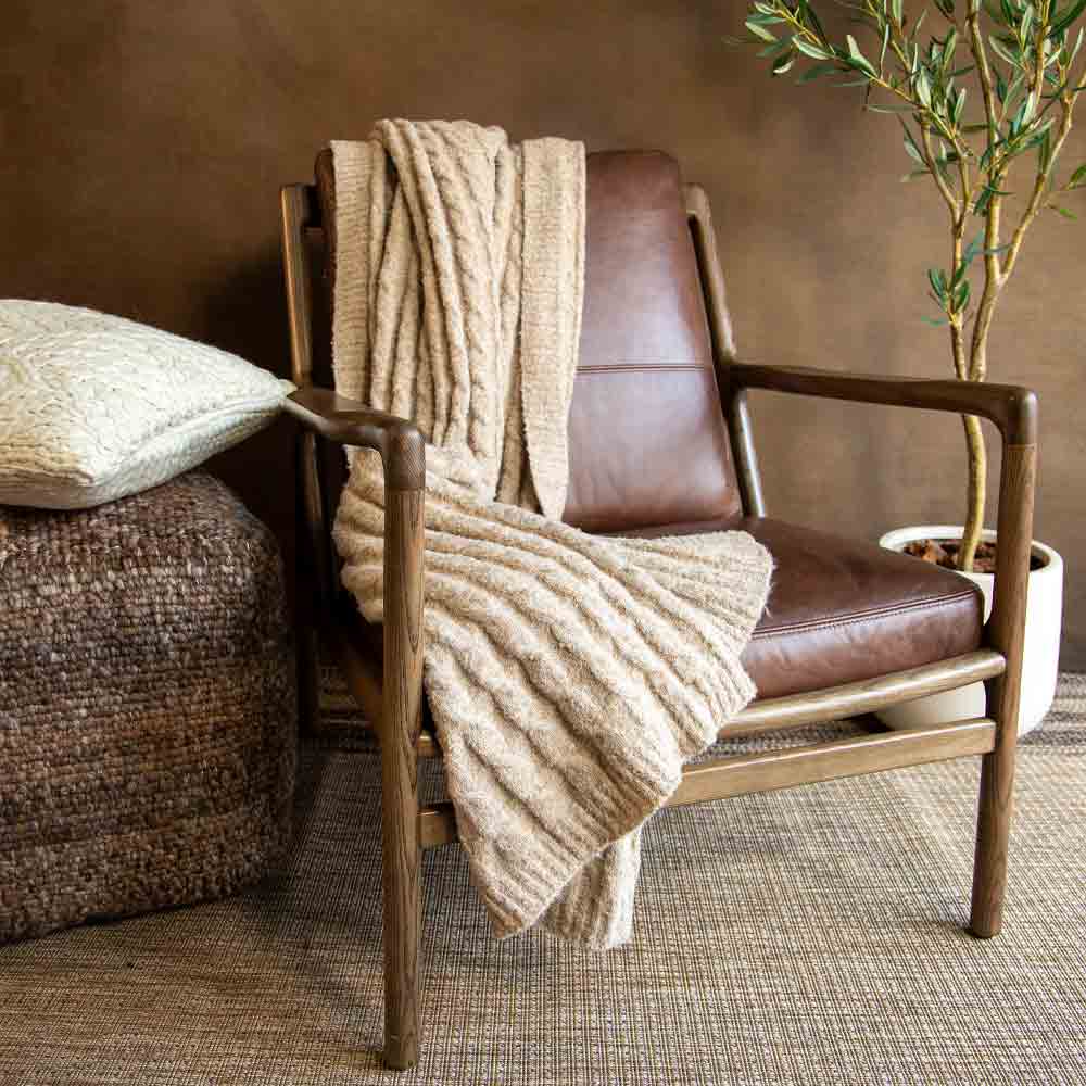 A mid-century modern wooden armchair with brown leather seating, styled with a chunky beige knit throw blanket draped over it, set in a cozy room with a textured brown wall, a woven pouf, and a potted olive tree.